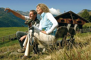 Pärchen sitz neben der Alm auf der Parkbank- der Mann deutet auf etwas in der Ferne und die Frau schaut mit dem Fernglas in der hand in diese Richtung