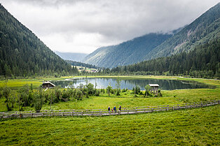 grünes Tal, leicht graue Wolken, wenig Sonnenschein- dennoch bwundert die kleine Wanderfamiilie auf den Wiesen den wunderschönen Bergsee