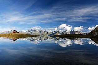 Frontalansicht einen glasklaren Bergsees- in ihm spiegeln sich die Berggipfel aus der Ferne und hinten Links im Bils bewundert man eine  große urige Alm