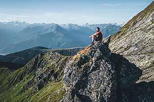 Hoch oben am Bergkamm sitzt ein Mann auf einem Felsen und betrachtet das traumhafte Panorama der unzähligen Berge und Gipfel im Hintergrund