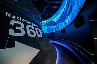 dunkle innenansich des Nationalparkgebäudes- weiße schrift auf schwarzen Steinplatten zeigen den Rundgang an- neonblau leuchtende 360grad Bildschirme die das Panorama der Berge zeigt