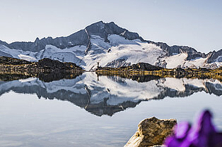 Flache Nahaufnahme einer Bergsees- im Hintergrund am anderen Ende des Ufers steht eine Person mit den Armen in der Luft und dem Berggipfel erstreckt sich hinter ihm