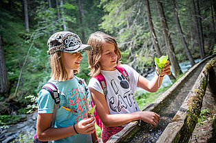 Im Wald beim Fluss, eine Wassertränke mit fließendem Bergwasser- zwei junge Mädchen benutzen gefaltete Blätter als Trinkbecher
