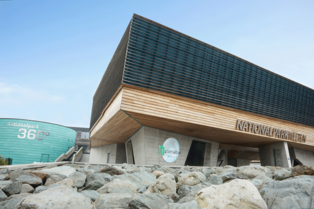 Aussenansicht des Nationalpark Gebäudes- unten sehr Breite Betonsäulen, dann eine Holzaufbau und darauf sehr hohe Fensterfront, geschützt mit dunklen holzpaneelen