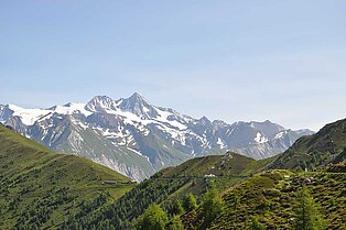 Im Vordergrund grüne Bergkämme und in er Ferne Berge die von grün über felsig brauch bis hin zu angezuckerten weißen Berggipfeln