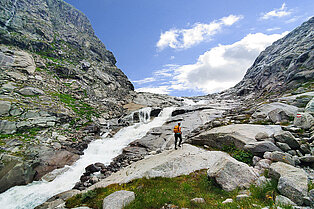 Links und rechts Felswändein der Mitte ein Fluss der ins Tal runter fließ, daneben ein Wanderer auf einem Felsbrocken