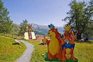 ein kleiner Kinderspielplatz mitten in den Bergen- Im Vordergrund ein Holzschild mit einem Murmeltier mit Schmetterling auf dem Kopf das den Spielplatzpräsentiert- ein weiters macht daneben einen handstand und ein Schmetterling fliegt über die Füße