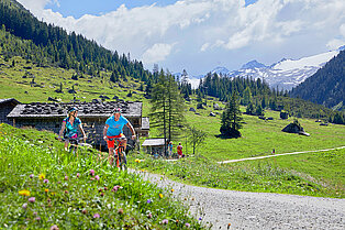 Bergpanorama- im Vordergrund ein radelndes Päarche fahren von ihrer kleinen Hütte weg- im Hintergrund felsige grünen Wiesen, Bäume und Berggipfel