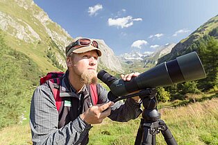 Nahaufnahme eines Wanderers mit Fernrohr auf Stativ- bereit die Natur unter die Lupe zu nehmen- im Hintergrund das Tal und die berge