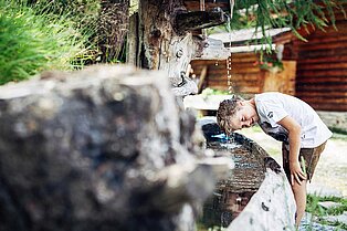 Kleiner blonder Bub in Lederhose hält seinen Kopf zur Erfrischung unter den Wasserstrahl beim Holzbrunnen vor der Almhütte