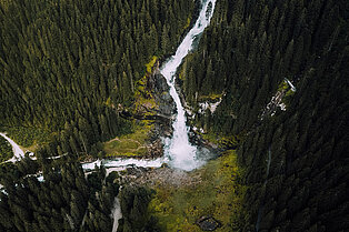 Ansicht von oben auf einen Wald, grüne Wiese und Felsen- in der Mitte bahnt sich ein straker fluss von oben in die Mitte und macht die Kurve nach links