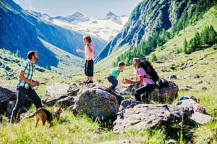 Im grünen tal stehen zwei Kinder auf Felsbrocken- Die Tochter springt Papa lachend in die Arme, der Sohn der Mama