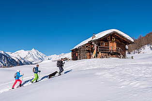 Schneelandschaft- Wanderguide und Hund begleitet ein Wanderpäarchen mit Schneeschuhen an einer Berghütte vorbei zum Gipfel