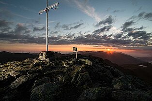 Abenddämmerung- Das Gipfelkreuz direkt vor sich hat man den Blick in die Ferne auf andere Berggipfel- Am Horizont sind ein paar schwarze Wölkchen und die Sonne die untergeht zu erkenne