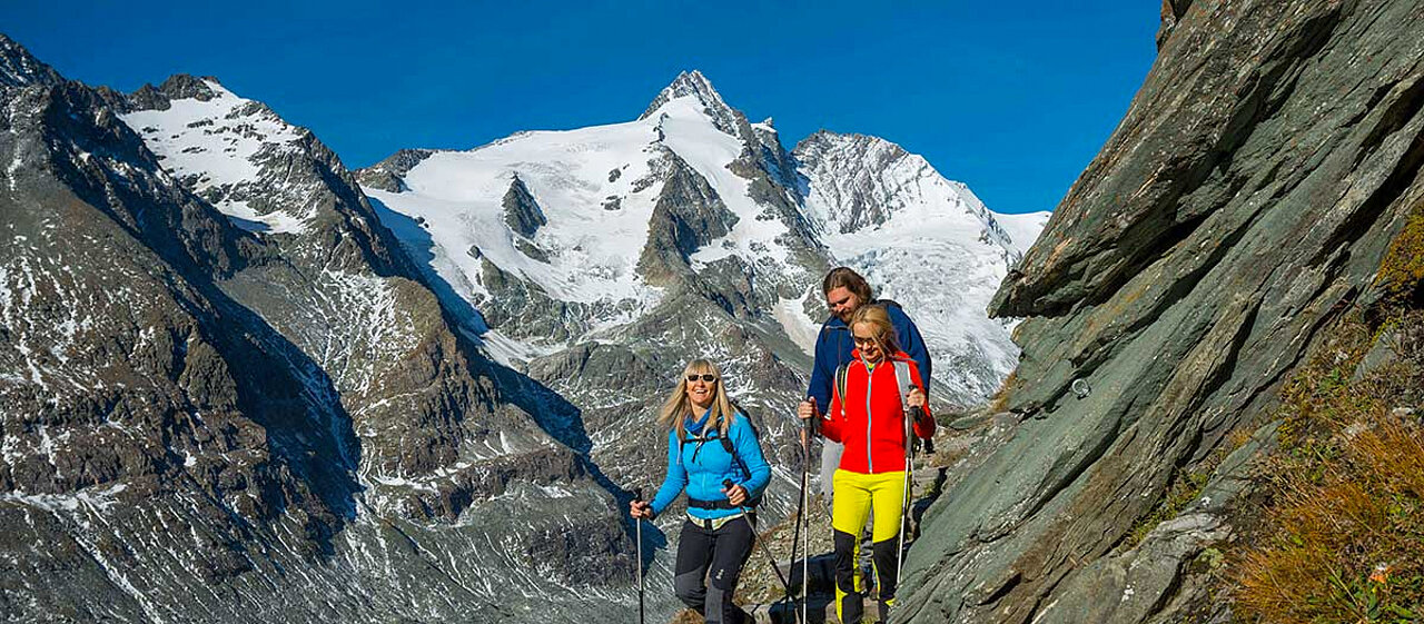 Mann und 2 Frauen gehen mit Wanderstöcken den Berg an der Kante herunter