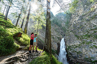 Mutter stehe mit ihrem Sohn im Arm im Wald auf dem Weg nach oben und bewundert den Wasserfall der sich aus der Felswand den Weg ins Freie bahnt