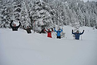 eine Gruppe Schneeschuhwanderer steht teilweise Hüfthoch im Schnee vor einem Wald- jubelnd für das Fotot reisen sie die Arme nach oben